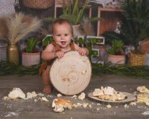 A baby with cake on their face and body kneels on the floor, holding a large wooden slab surrounded by scattered pieces of cake. There are plants in the background.
