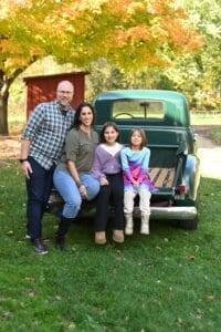 A family of four poses in front of a green vintage truck, with trees and a red shed in the background on a sunny day.