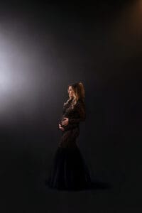 A pregnant woman in a long, black lace dress stands in profile against a dark background, holding her belly with both hands.