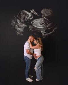 A couple stands closely together, with the woman’s hand on her pregnant belly, in front of a large ultrasound image projected behind them.
