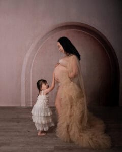 A pregnant woman in a beige tulle gown stands sideways as a young girl in a white dress gently touches her belly.