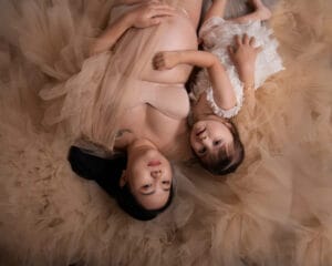 A pregnant woman in a nude tulle dress lies on the floor beside a young girl in a white dress, both looking up at the camera.