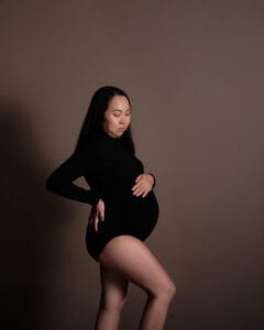 A pregnant woman in a black long-sleeve bodysuit stands against a plain brown background, with one hand on her lower back and the other on her stomach.