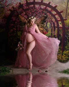 A pregnant woman wearing fairy wings, a pink sheer gown, and a flower crown stands barefoot by a pond, holding flowers, with a floral garden arch and pink blossoms in the background.