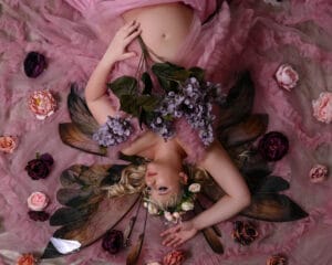 A person wearing a pink dress and floral crown lies on pink fabric surrounded by flowers, holding purple blooms, with translucent wings visible behind them.