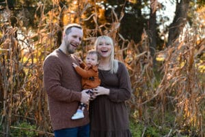 A smiling man and woman stand outdoors in front of tall dried plants, holding a small child dressed in autumn clothing.
