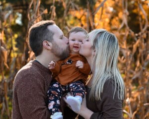 A man and woman kiss a smiling baby on the cheeks while holding the baby outdoors in front of dried corn stalks.
