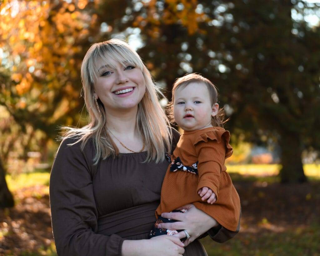 A woman with blonde hair holds a toddler in an autumn park, both wearing brown outfits. Trees with orange leaves are visible in the background.