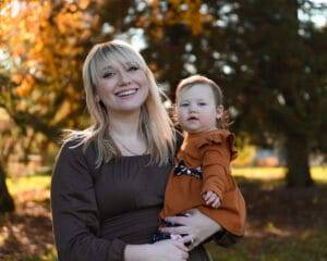 A woman with blonde hair holds a toddler in an autumn park, both wearing brown outfits. Trees with orange leaves are visible in the background.