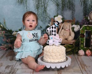 A baby in a blue dress sits on a wooden floor next to a white frosted cake with a “ONE” topper, surrounded by flowers, greenery, and a plush teddy bear.