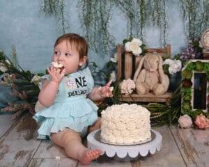 A baby in a blue dress sits on the floor eating a white frosted cake, surrounded by flowers, a teddy rabbit, and a decorative "ONE" sign.