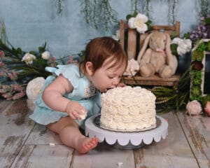 A baby in a light blue dress leans forward to eat a white frosted cake with their mouth. There are flowers and a stuffed bunny in the background.