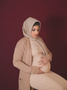 A pregnant woman wearing a beige hijab and matching outfit sits on a stool, holding her baby bump against a maroon background.
