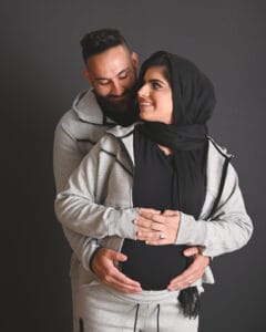 A smiling couple poses together; the man stands behind the woman, both hold her pregnant belly. They wear matching grey outfits, and the woman has a black hijab.