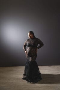 A pregnant woman in a long, sheer black lace dress stands on a wooden floor against a dark gradient background, looking down and holding her belly.