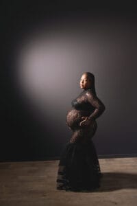 Pregnant woman in a black lace dress stands with one hand on her belly and the other on her hip against a dark background.