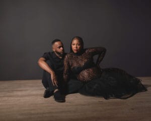 A man in black sits on a wooden floor next to a woman in a sheer black lace dress, who is posed with one hand on the ground against a dark background.