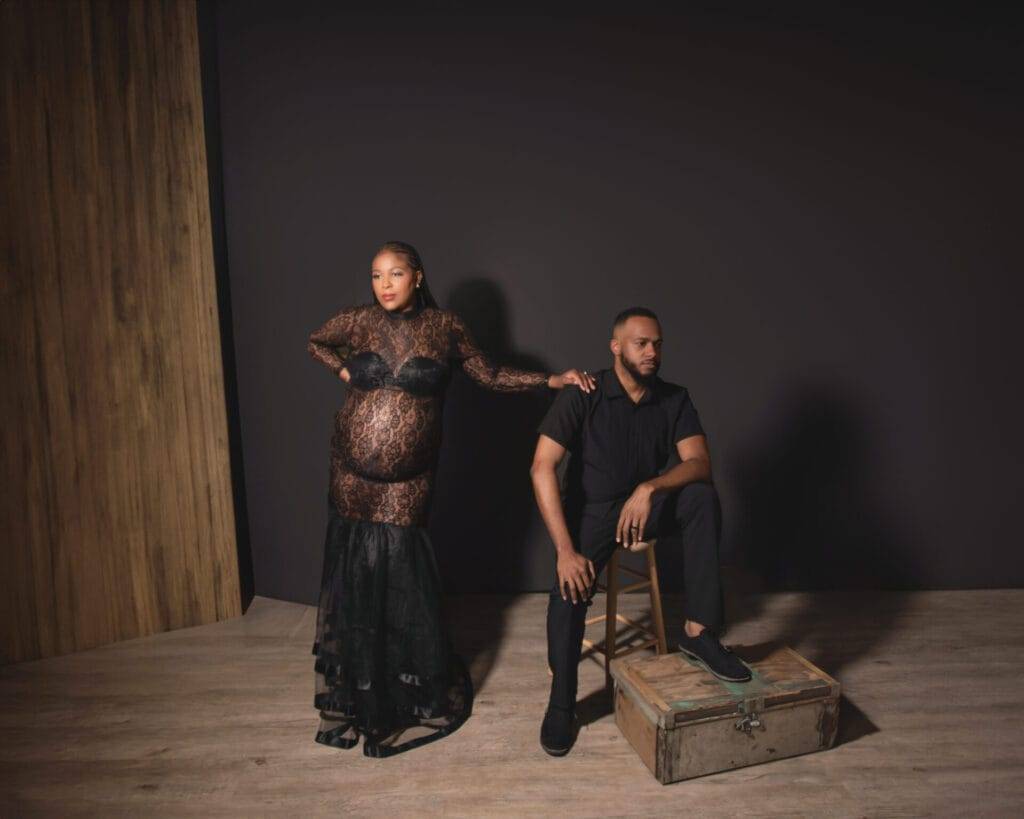 A pregnant woman in a black lace dress stands beside a seated man dressed in black, against a dark backdrop with a wooden floor and wall.