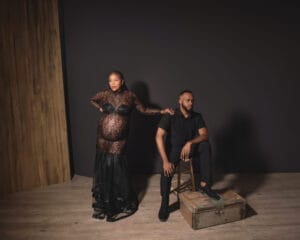 A pregnant woman in a black lace dress stands beside a seated man dressed in black, against a dark backdrop with a wooden floor and wall.