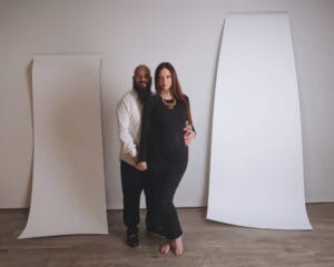 A man and a pregnant woman stand together in a studio, posing between two large curved white panels on a wooden floor.