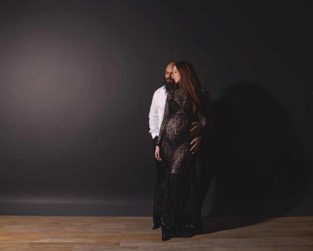A couple poses together in front of a dark background; the woman wears a black lace dress and appears pregnant, while the man stands behind her in a white shirt.