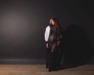 A couple poses together in front of a dark background; the woman wears a black lace dress and appears pregnant, while the man stands behind her in a white shirt.