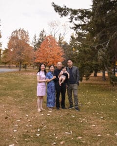 Five people, including a baby, stand together in a park with autumn trees and fallen leaves in the background.