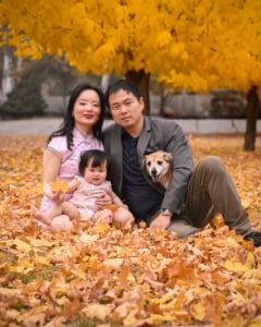 A family with a baby and a dog sits on fallen autumn leaves under a yellow-leaved tree, posing for a portrait outdoors.