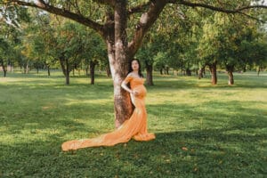 Pregnant woman in a long flowing orange dress stands by a tree in a sunlit park with green grass and trees in the background.