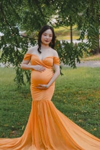 A pregnant woman in an orange off-shoulder gown stands on grass under a tree, holding her belly with both hands.