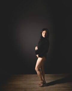 Pregnant woman in a black bodysuit and heels stands on a wooden floor against a dark background, holding her belly and looking at the camera.