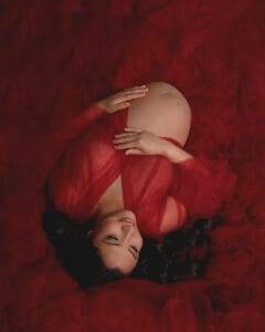 Pregnant woman lies on her back in a sheer red gown, hands resting on her bare belly, surrounded by red fabric.