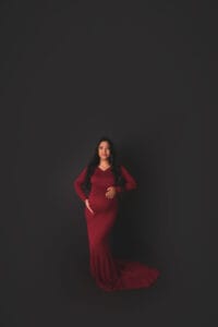 A pregnant woman in a long, fitted red dress stands against a plain, dark background, resting one hand on her belly and the other on her hip.
