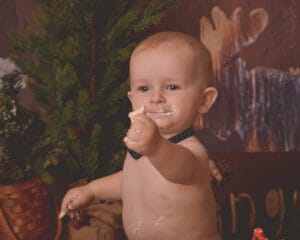 A baby with light hair, wearing a black polka dot bow tie, sits shirtless, holding cake with some smeared on their face, in front of rustic decor and a small pine tree.