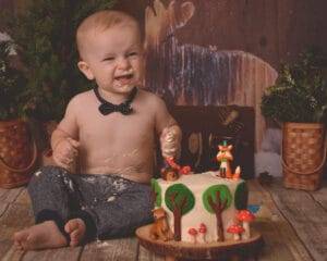 A smiling baby with a bow tie sits shirtless on a wooden floor, surrounded by cake, with a woodland-themed cake in front and rustic decor in the background.