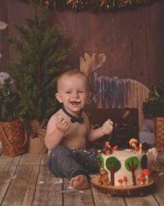 A smiling toddler with a bow tie sits on a wooden floor, covered in cake, with a decorated woodland-themed cake and rustic forest decor in the background.