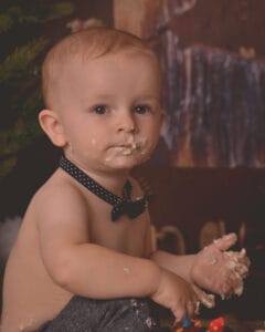A baby wearing a black polka dot bow tie has cake or icing smeared on their face and hands, looking at the camera.