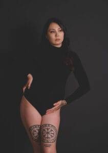 A person in a black long-sleeve bodysuit poses against a dark background, showing tattoos on their thighs and hand.