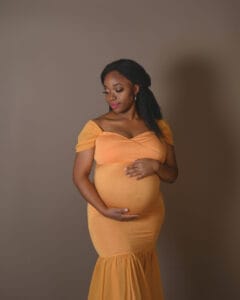 A pregnant woman in an off-the-shoulder yellow dress stands against a plain brown background, gently cradling her belly with both hands.