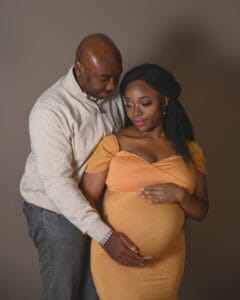 A man stands behind a pregnant woman in an orange dress, both holding her baby bump and looking down with gentle expressions, against a plain background.