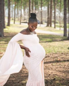 A pregnant woman in a light pink off-shoulder dress stands outdoors in a wooded area, holding her belly and smiling.