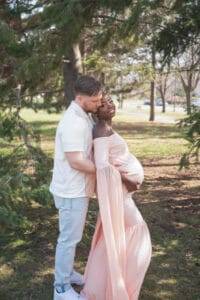 A man stands behind a pregnant woman in a long pink dress, embracing her and kissing her forehead in a park setting with trees and sunlight.