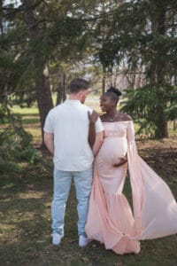 A couple stands outdoors on grass; the woman in a long, flowing pink dress holds her pregnant belly and looks at the man, who is wearing light pants and a white shirt. Trees are in the background.