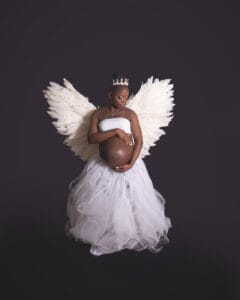 Pregnant woman wearing a white tutu skirt, white angel wings, and a crown, standing against a dark background and cradling her belly.