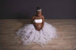 A pregnant woman wearing a white tulle skirt and a crown sits on a wooden floor, gently holding her bare belly and looking down.