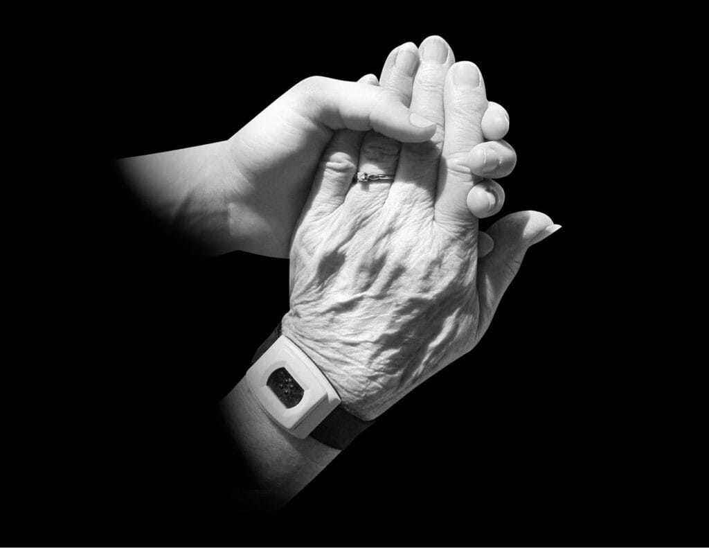 A younger hand gently holds an older hand wearing a ring and a medical alert wristband, against a black background.