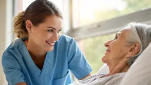 A nurse in blue scrubs smiles and leans toward an elderly woman lying in a hospital bed by a window.