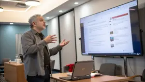 A man stands beside a large screen displaying a presentation as he gestures with his hands in a conference room.