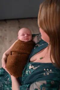 A woman in a green dress holds a sleeping newborn wrapped in a brown knitted blanket, viewed from above and slightly behind her shoulder.