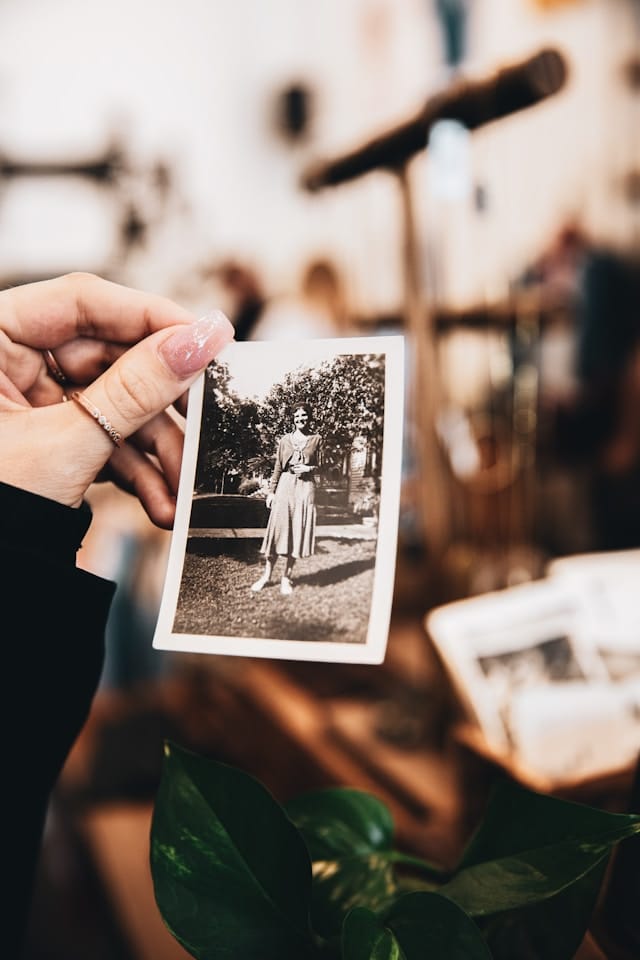 A hand with manicured nails holds a black-and-white photo of a woman standing outside, with blurred objects and plants in the background.